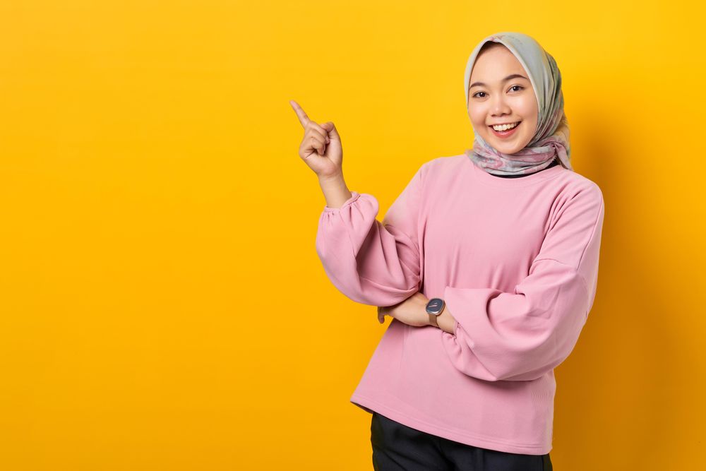 Happy Muslim woman in scarf and pink sweater pointing up at copy space studio shoot