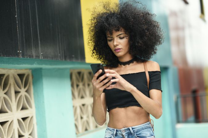 Young woman with afro hair checking phone near a modern colorful building