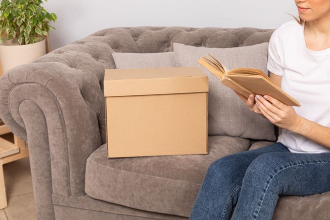 A woman with a box is sitting on the sofa reading a book