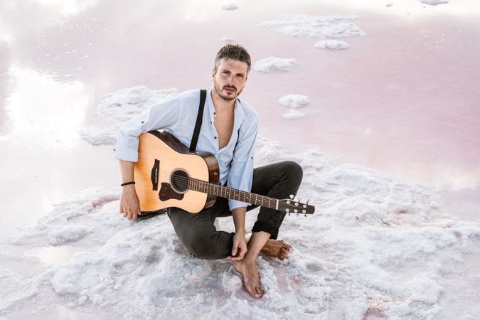 Dreamy musician resting with guitar on sand surrounded with water reflecting colorful pink sky