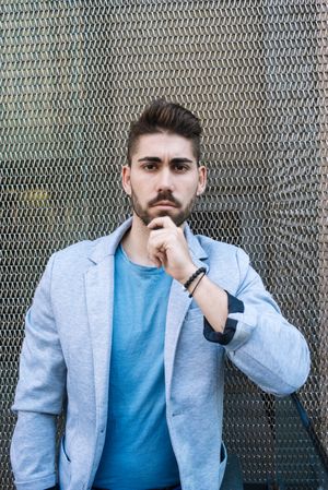 Portrait of young bearded man leaning on wired wall and looking at camera, vertical
