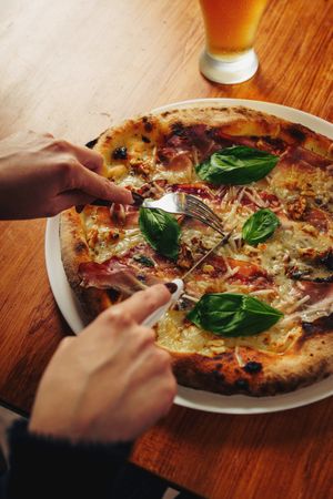 Person slicing fresh basil pizza with fork and knife at table