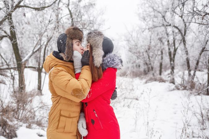 Teenage boy and girl standing in snowy forest in winter coats and fur hat and kissing