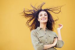 Female in army green jumpsuit smiling in front of mustard wall with arms folded & hair blowing back