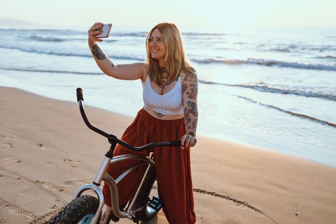 Cheerful woman taking selfie on smartphone while resting with bicycle at sunny shore