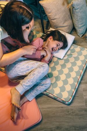 Happy mother tickling to young daughter while laughing lying over mats in night pajama party