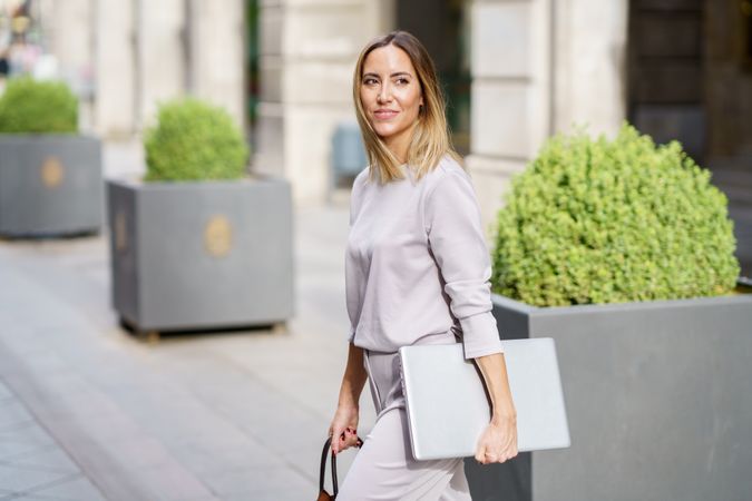 Smiling businesswoman with laptop crossing road in city modern district