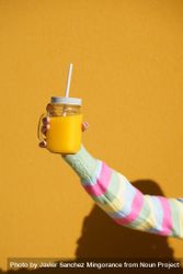 Vertical shot of person in striped sleeve holding cup of orange juice on yellow wall 5l8n65