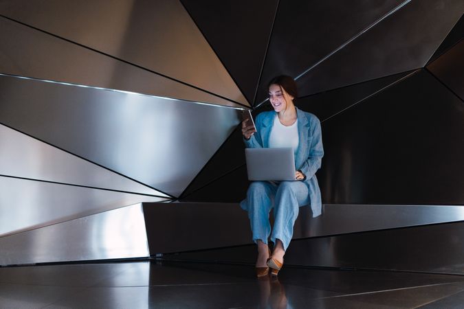 Cheerful woman with laptop browsing smartphone in futuristic studio