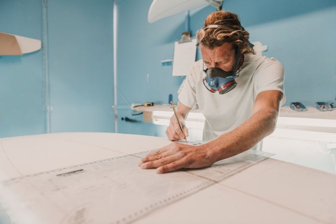 Man in respirator measuring surf board in workshop