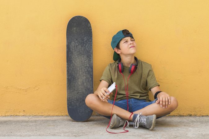 Front view of a teenage boy sitting on ground with skateboard leaning on a yellow wall while holding a mobile phone