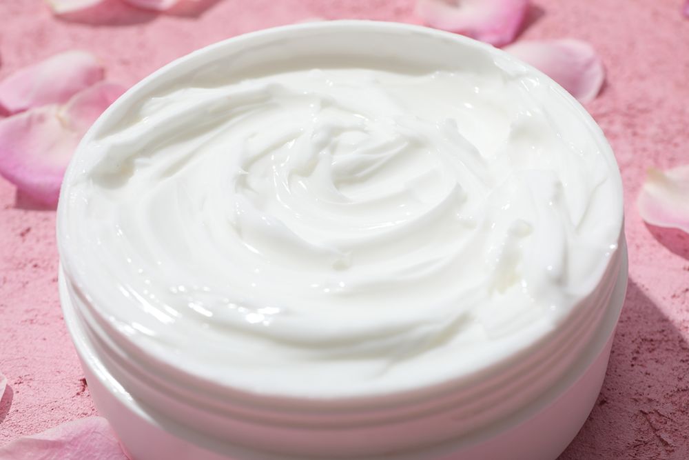 Jar with cream and rose petals on pink background, close up