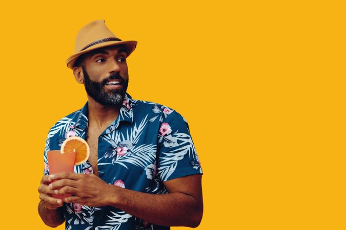 Male holding a cocktail while wearing colorful shirt with yellow background, copy space