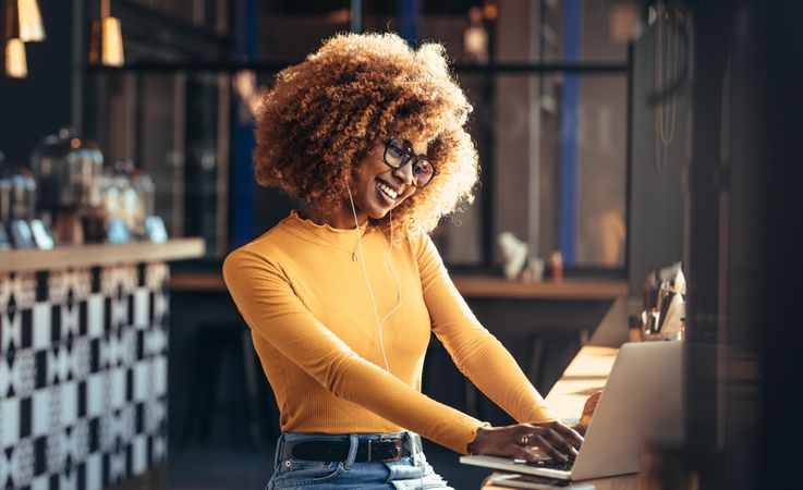 Cheerful young Black woman with curly hair and eyeglasses working on her laptop
