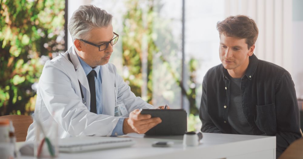 Doctor with Stylish Gray Hair Showing Analysis Results on Tablet Computer to a Healthy Young Man During Visit to a Health Clinic.
