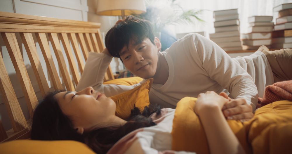 Young Korean Couple Sleeping in Bed, Sun Shines on Them, They Look at Each Other and Greet New Sunny Day.