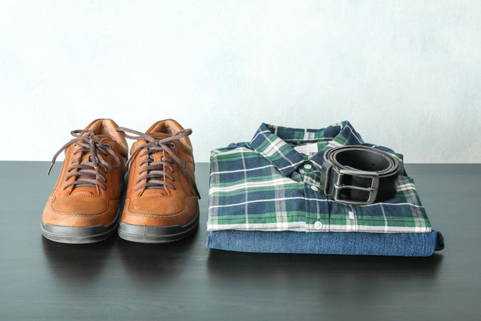 Stack of shirt, jeans and belt with shoes on table against light background, space for text