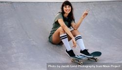 Woman making peace sign sitting on ramp with skateboard 56e1V0