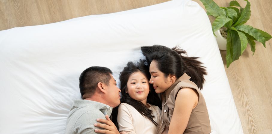 Top view of Asian family lying on bed. Parent kissing daughter cheek