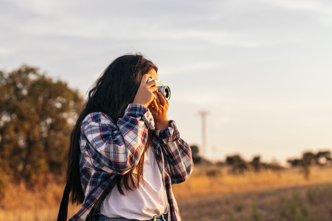 Young woman taking photos with analog camera in the field at sunset