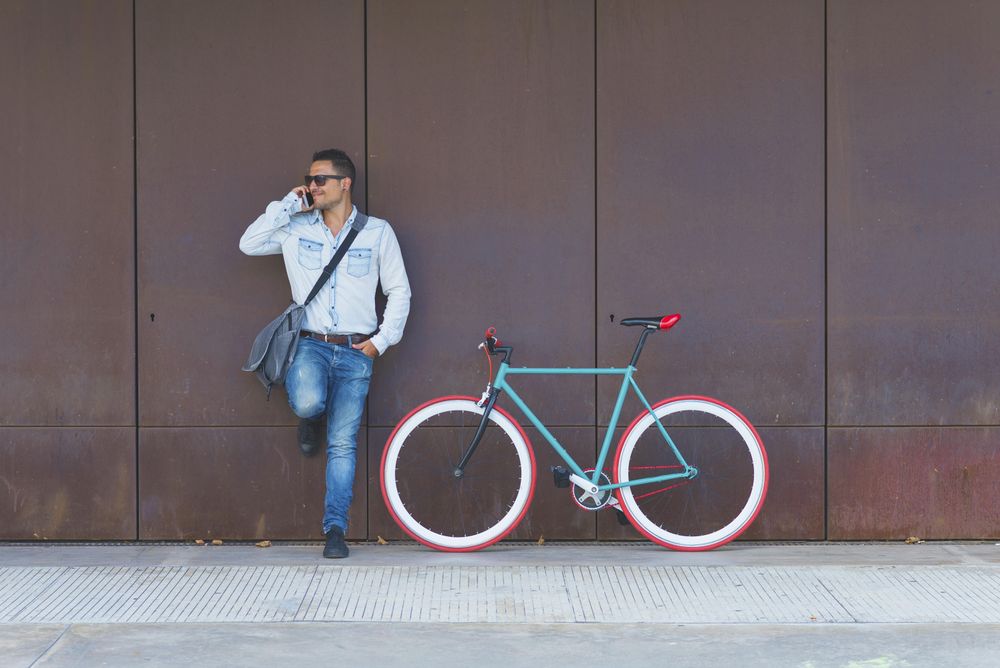 Male in sunglasses standing with red and green bicycle leaning on wall and talking on phone