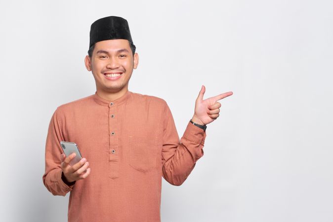 Happy Muslim man in kufi hat holding mobile phone and pointing to the side