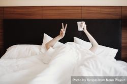 Person under light bed sheets holding ceramic mug and making peace sign 4Z7lNb
