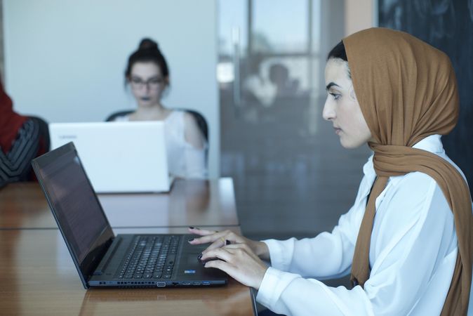 Woman wearing hijab working on her laptop in office with two other women