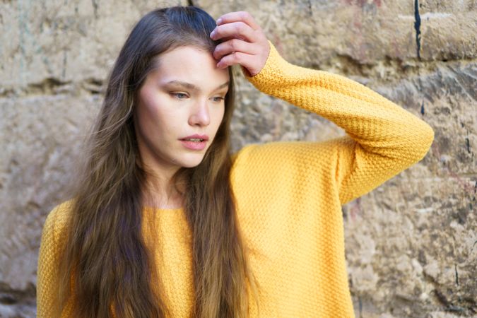 A contemplative woman dressed in a bright yellow sweater poses against a textured stone wall