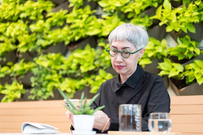 Trendy Asian woman with green frames sitting in cafe looking at phone