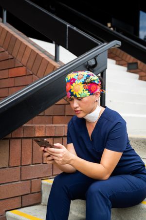 Nurse in head covering and scrubs sits on stairs outside checking phone on break