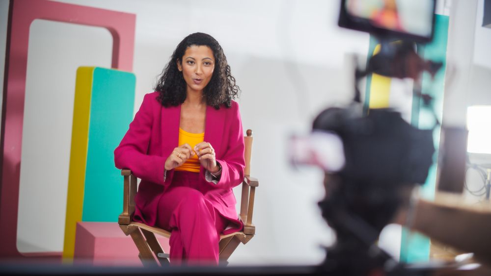 Young Female Host In Vibrant Pink Suit Engaged In Lively Discussion On Colorful Set, Captured On Camera For A Modern Talk Show, Emphasizing Dynamic Storytelling And Visual Appeal.