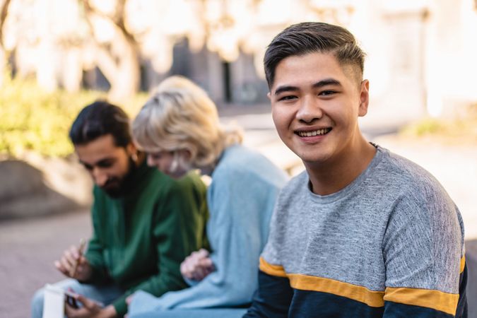 Portrait of a cheerful Asian male student with friends with a blur background, enjoying an outdoor gathering