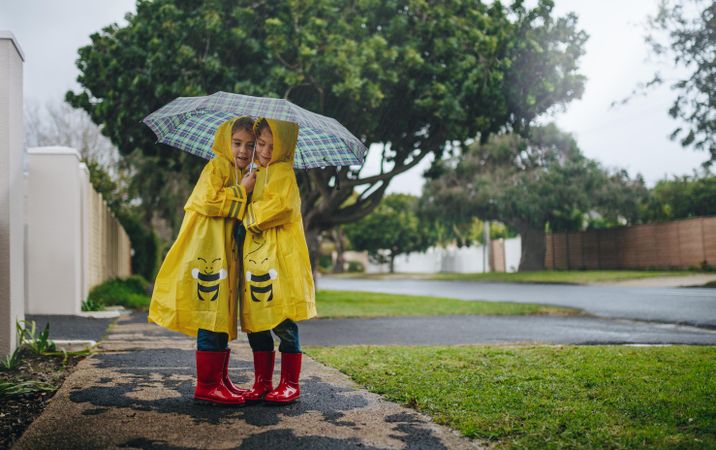Twin sisters in waterproof coats standing together under umbrella outside