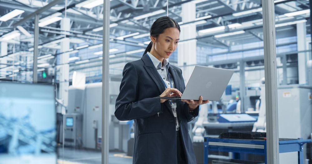 Asian Factory Manager Working on Laptop at Futuristic Industrial Manufacture with Autonomous Robotic Assembly Line. Specialist Monitoring Production, Planning Work for Employees