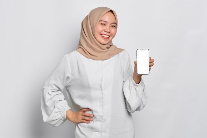 Happy Asian Muslim woman in a bright studio shoot presenting cell phone with mockup screen
