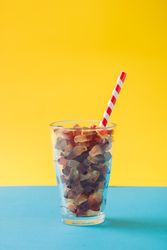 Glass full of gummy candies with straw on blue and yellow background