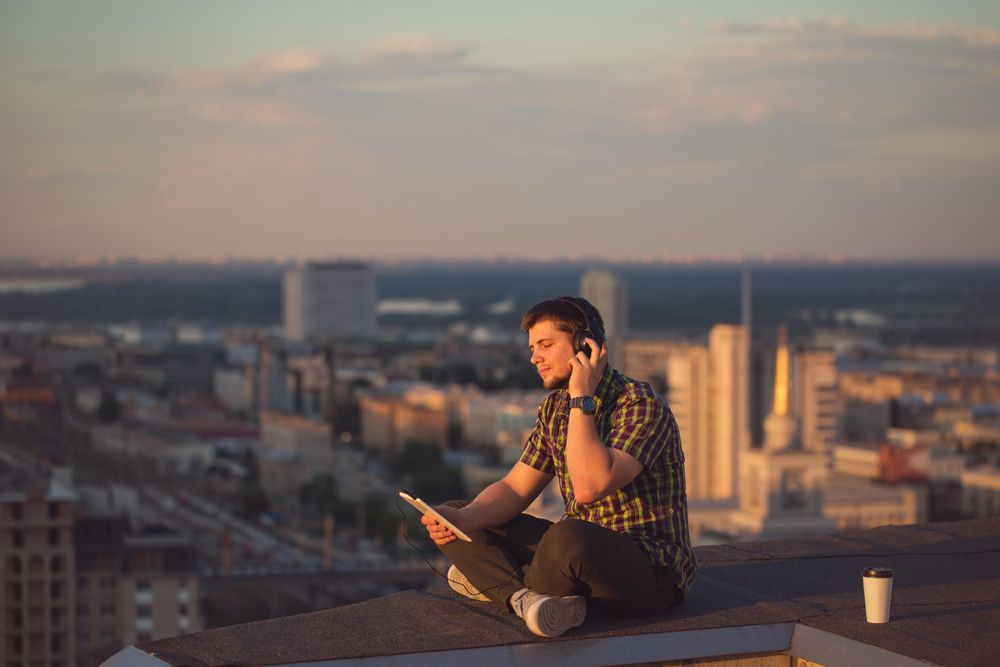 Male using digital tablet with headphones with takeaway coffee cup on roof over skyline