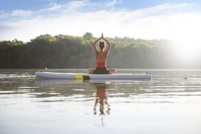 Rearview of woman sitting on paddleboard with arms up on lake