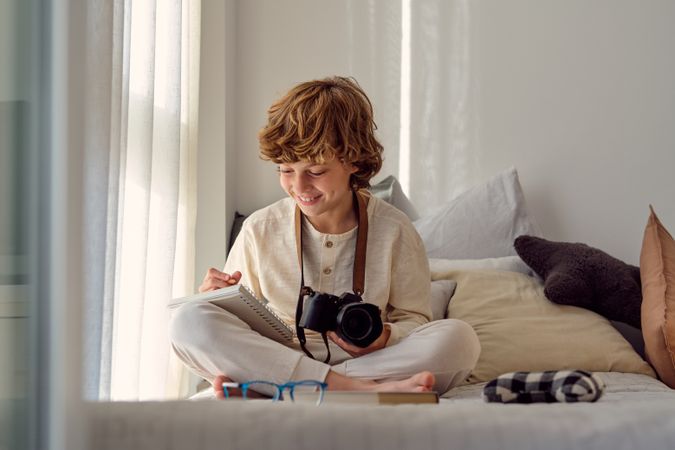Cheerful boy with photo camera writing in copybook on bed