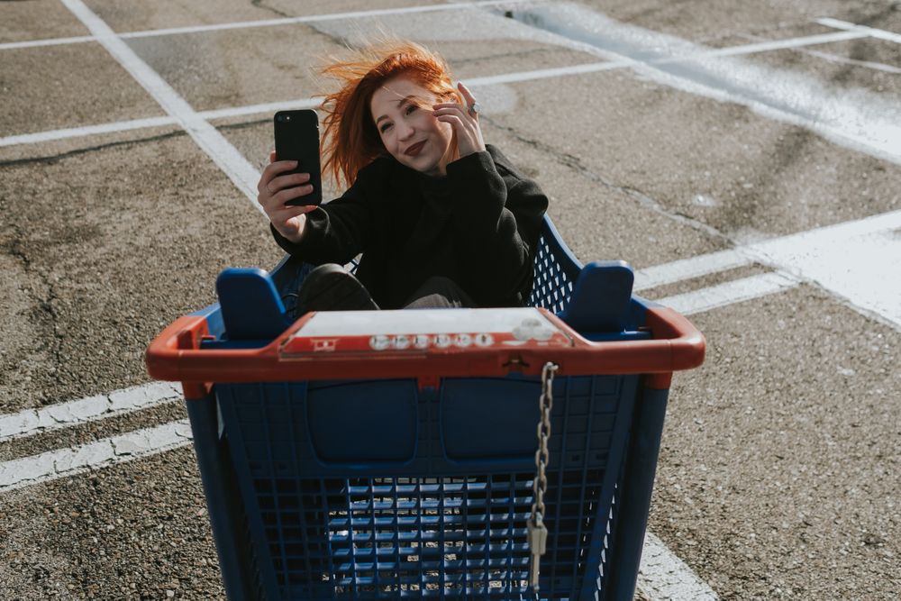Smiling woman with smartphone in shopping trolley in parking lot