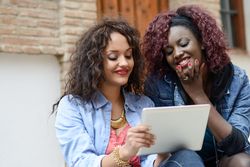 Black female friends reading something together on a tablet while sitting outside