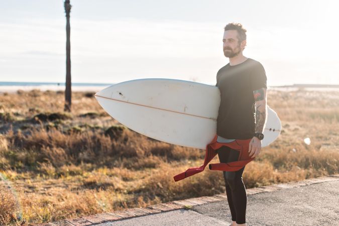 Tattooed man standing on road near beach holding surfboard