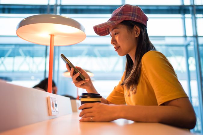 Asian woman browsing smartphone while drinking coffee at table under lighting lamp