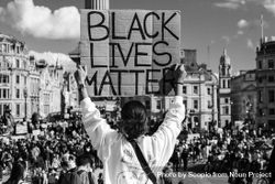 Grayscale back view of woman holding a banner that says " Black Lives Matter" against a crowd in London 49YxL4