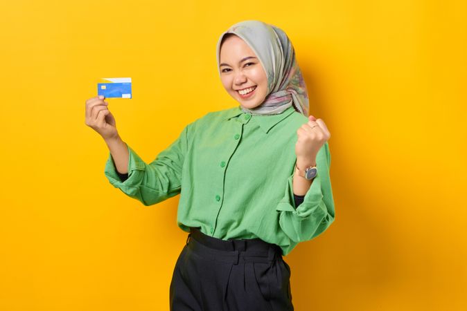 Muslim woman in headscarf and green blouse holding her credit card and clenching fist in celebration