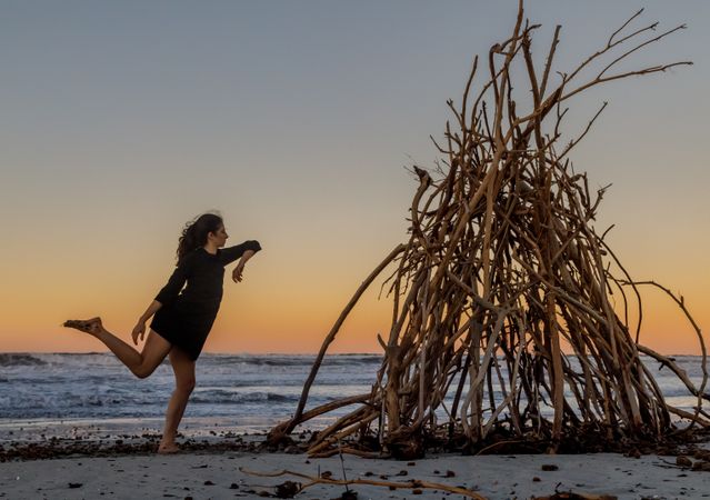 Young woman dancing on shore near water and wooden twigs placed for campfire