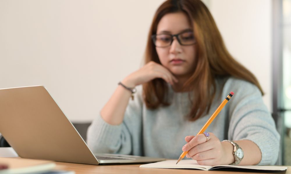 A woman wearing glasses uses a pencil to write on a notebook with a laptop on the table.