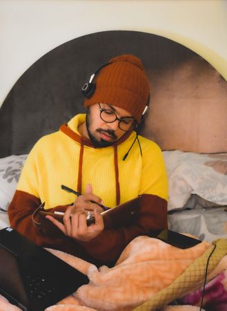 Man in yellow hoodie using smartphone writing notes and using laptop in bed
