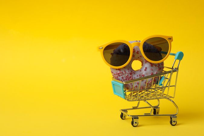 Shop trolley with sunglasses and donut on yellow background